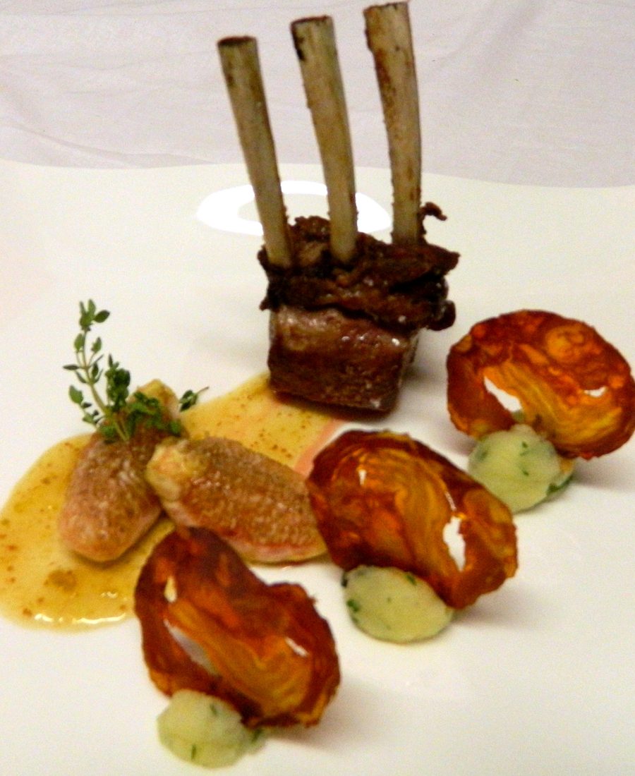 Costolette di agnello impilate su un piatto quadrato bianco, con chips di verdura e quenelle di purea