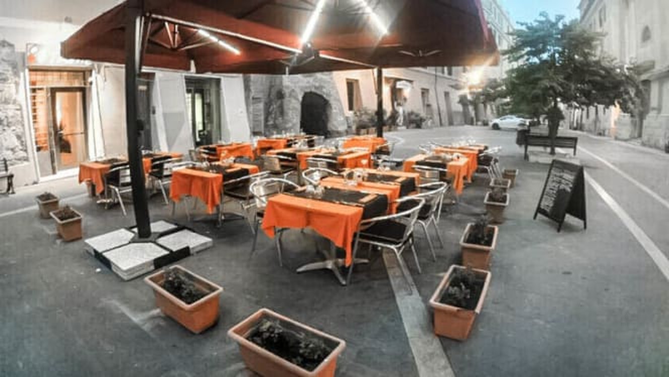 I tavoli esterni di 80 Fame sulla piazza pedonale, con tovaglie arancioni, ombrelloni aperti e vasi di piante intorno
